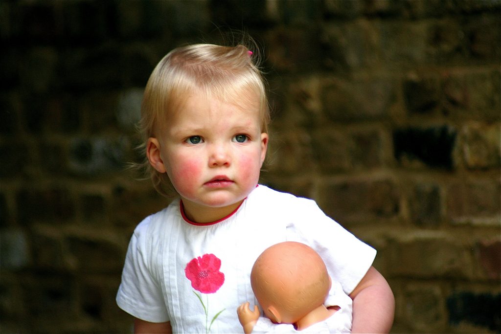 Pretty blonde girl holding doll and wearing white dress with red flower
