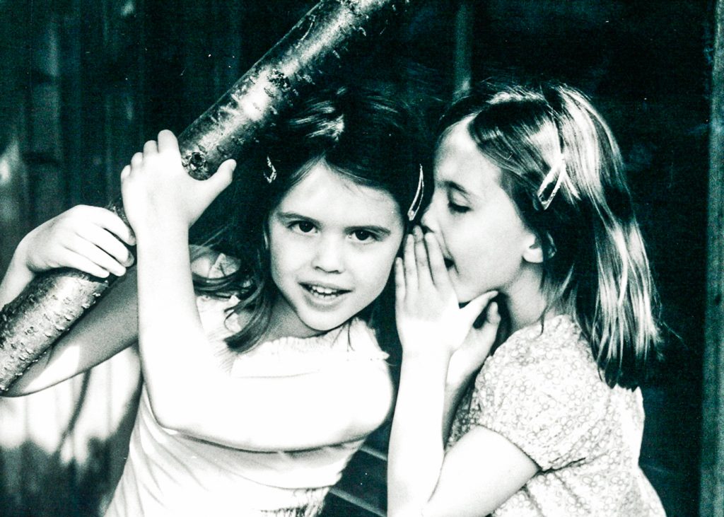 black and white photo of a little girl whispering to her friend outside in a garden