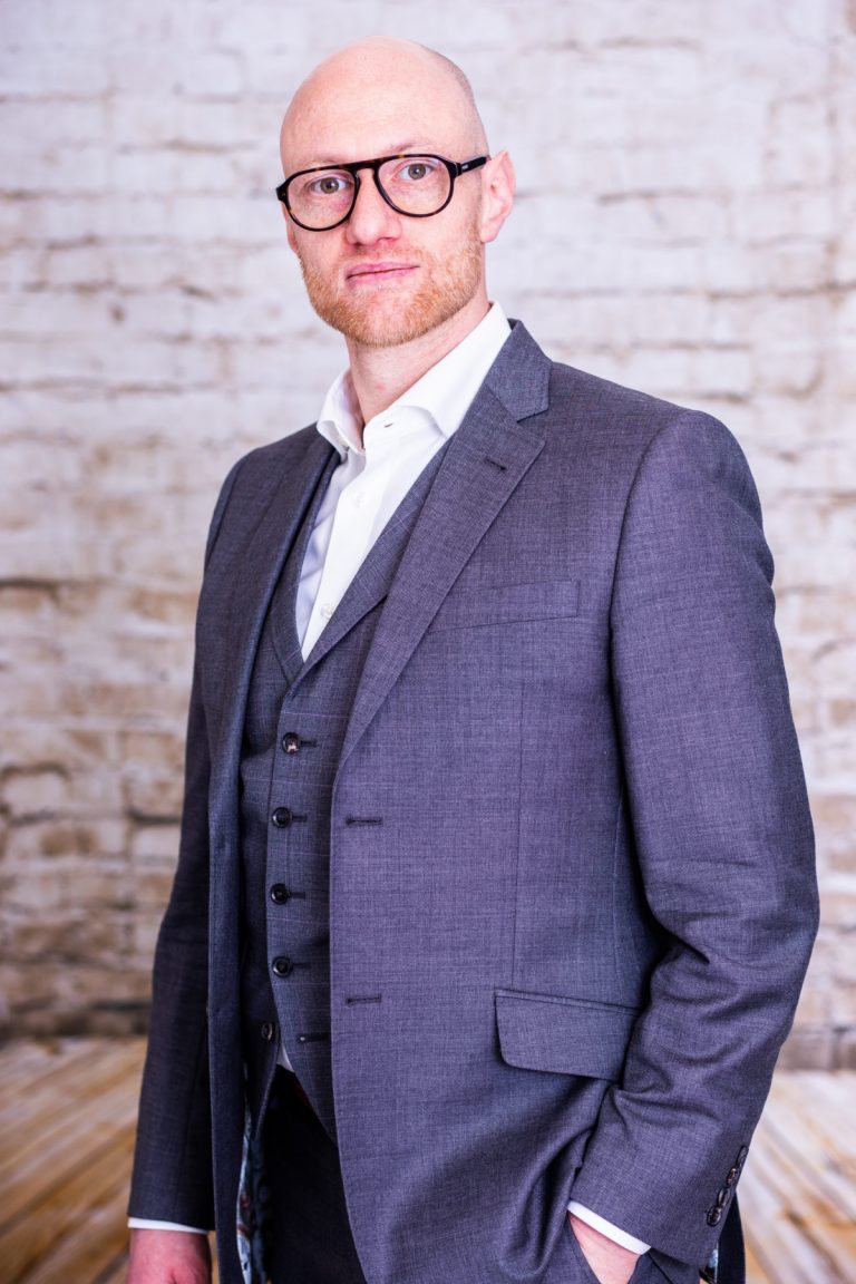 smart bald man with beard in suit against white brick wall